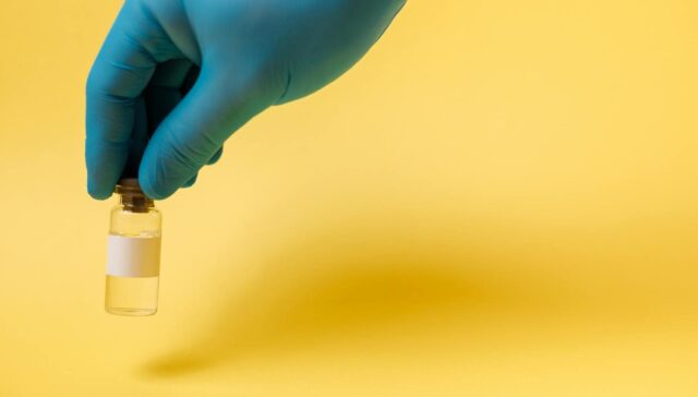 Close-up of a blue gloved hand holding a vaccine vial on a yellow background.