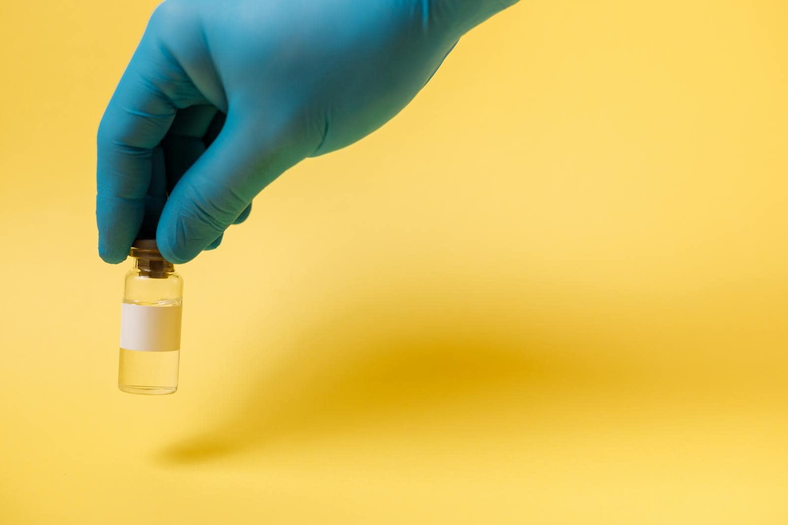 Close-up of a blue gloved hand holding a vaccine vial on a yellow background.